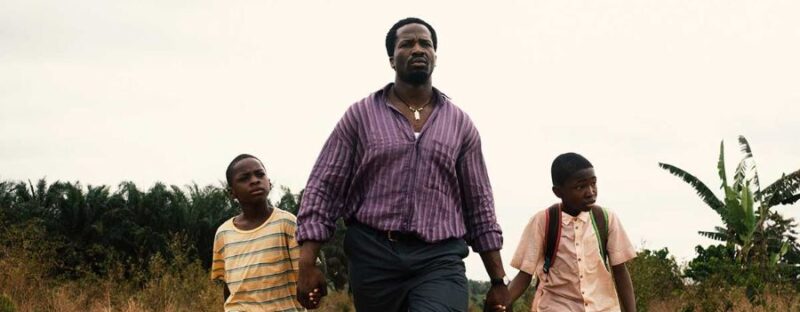 Folarin, homem negro, vestindo camisa roxa e calça escura, caminha por uma estrada de terra segurando a mão de dois menino, enquanto outro o da esquerda usa uma camiseta amarela com listras azuis e o da direita usa uma camisa clara de tom alaranjado. Ao redor, a vegetação e o céu claro compõem a paisagem.