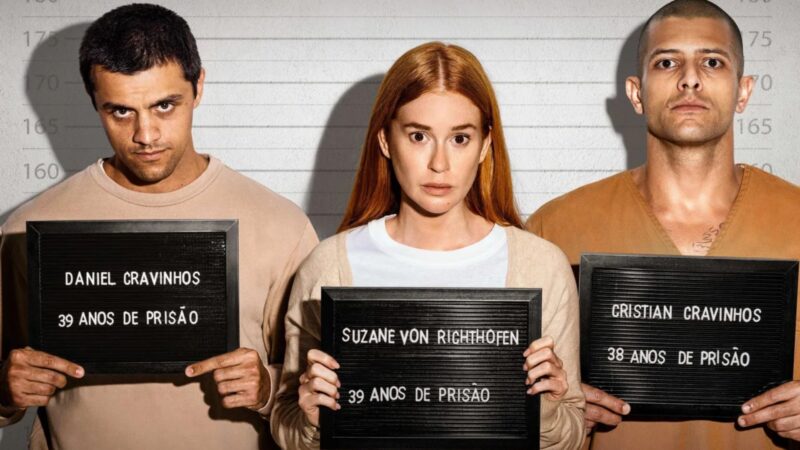 [Texto alternativo: Cena promocional de Tremembé. Três personagens posam para fotos de ficha criminal, segurando placas com seus nomes e penas: Daniel Cravinhos, Suzane von Richthofen e Cristian Cravinhos. O fundo branco com marcações de altura remete ao registro policial, evidenciando o aspecto realista e documental da série.]