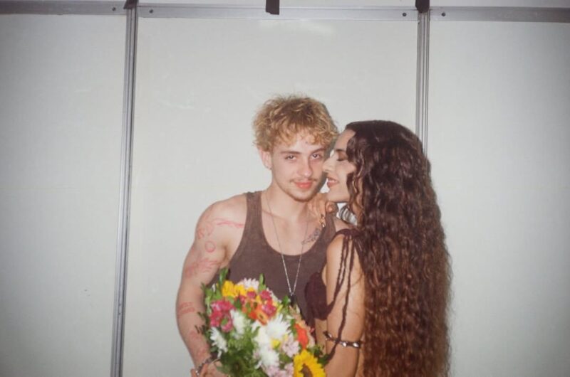 Foto de Marina Sena ao lado de Juliano Floss que segura um buquê de flores. Marina está de perfil, sorrindo, com seus longos cabelos cacheados em destaque.