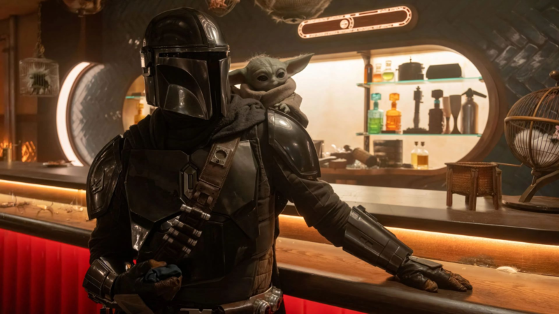 Cena de O Mandaloriano e Grogu ambientada em uma cantina futurista com iluminação quente em tons alaranjados e avermelhados. Em primeiro plano, o Mandaloriano aparece em pé apoiado no balcão de madeira, usando sua armadura metálica preta e prateada com acabamento brilhante e capacete refletivo que cobre completamente o rosto. Uma faixa marrom atravessa seu peito com pequenos compartimentos metálicos presos ao traje. Sobre seu ombro direito está Grogu, pequena criatura verde de grandes orelhas pontudas e olhos escuros expressivos, envolta em um casaco grosso bege de tecido felpudo. Ao fundo, prateleiras iluminadas exibem frascos coloridos e objetos futuristas, enquanto o ambiente curvo da cantina reforça a estética clássica de Star Wars.