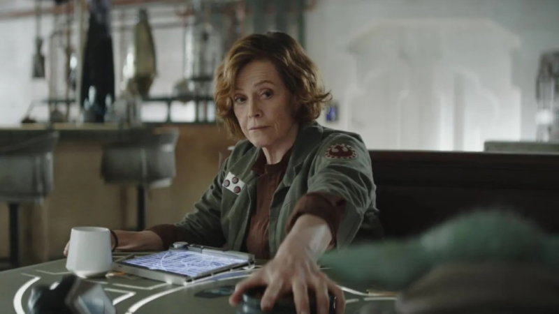 Cena de O Mandaloriano e Grogu mostrando a Coronel Ward, interpretada por Sigourney Weaver, sentada diante de uma mesa futurista iluminada suavemente. A personagem é uma mulher branca de cabelos curtos castanho-avermelhados e expressão séria e analítica. Ela veste um uniforme militar verde-oliva com detalhes e insígnias inspirados na Nova República. Com uma das mãos apoiada sobre a mesa tecnológica e a outra próxima de uma caneca branca, Ward observa alguém fora de quadro. Ao fundo, o ambiente apresenta paredes claras, estruturas metálicas e iluminação suave que reforçam a estética sci-fi do universo Star Wars.
