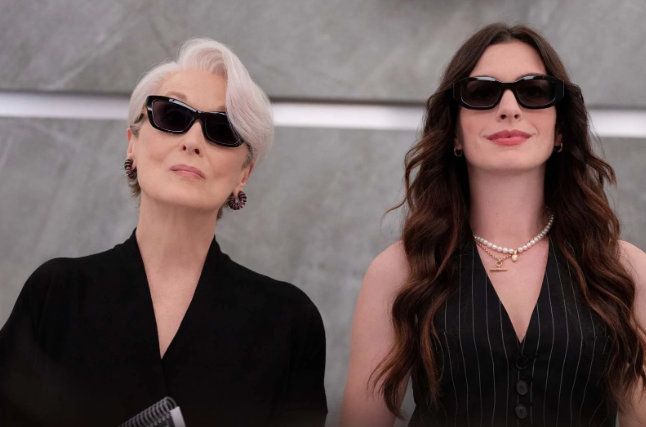 Duas mulheres estão lado a lado, encarando a câmera com expressão confiante. Ambas usam óculos escuros pretos e roupas em tons escuros. À esquerda, uma mulher mais velha, de cabelo curto grisalho, veste um blazer preto e brincos discretos. À direita, uma mulher mais jovem, com cabelo longo castanho ondulado, usa um colete preto com listras finas e um colar de pérolas. 