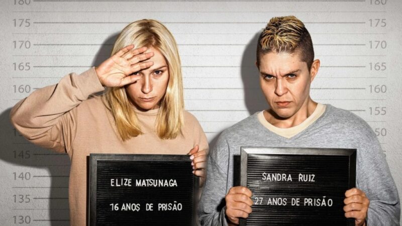 [Texto alternativo: Cena de Tremembé mostrando Elize Matsunaga e Sandra Ruiz posando para fichas criminais. Ambas seguram placas com seus nomes e penas. O olhar firme das personagens reforça a tensão entre arrependimento e frieza, um dos temas centrais da narrativa.]