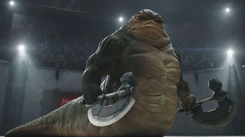 Imagem promocional de O Mandaloriano e Grogu destacando Rotta, o Hutt, em uma arena escura iluminada por refletores brancos. A criatura alienígena gigante possui corpo musculoso, pele enrugada em tons de verde e marrom e expressão séria e intimidadora. Rotta segura duas armas futuristas semelhantes a machados metálicos, uma em cada mão, enquanto encara a câmera de frente. Ao fundo, arquibancadas lotadas aparecem desfocadas sob a iluminação dramática do ambiente. No canto superior esquerdo, o selo branco ‘Empire Exclusive’ está sobreposto à imagem.