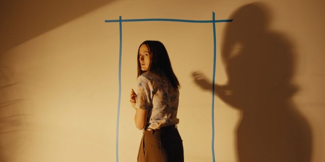 Cena do filme Backrooms: Um Não-LugarNa imagem, a personagem Mary está centralizada, de frente para uma parede branca com o desenho de uma porta em azul. O ambiente é iluminado por uma luz amarelada, provocando a sombra de Mary no canto direito. Mary é uma mulher branca, na faixa dos 40 anos, de cabelos escuros na altura dos ombros. Ela veste uma camisa florida de manga curta e uma calça marrom. Ela está olhando para trás com expressão de preocupação.