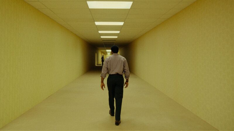 Cena do filme Backrooms: Um Não-LugarNa imagem, Clark está de costas para a câmera enquanto caminha em um corredor de paredes amarelas e luzes fracas. À sua frente há um trecho escuro com uma silhueta parada. Clark é um homem negro, na faixa dos 50 anos, de cabelos crespos, curtos e escuros. Ele veste uma camisa social rosa, calça preta e sapatos de couro.
