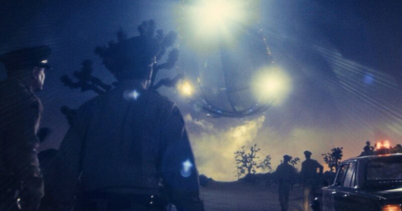 Cena do filme Dia DNa imagem, quatro policiais, de costas para a câmera, olham um OVNI no céu. Dois deles estão no canto esquerdo da foto enquanto os outros dois no lado direito, distantes da câmera, ao lado de uma viatura policial.. O cenário é noturno e a espaçonave é grande com fortes luzes acesas.