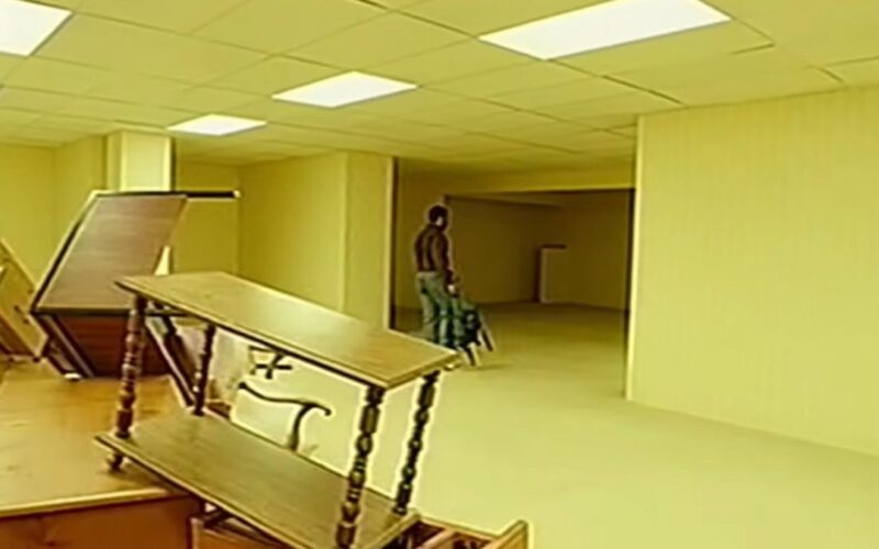 Cena do filme Backrooms: Um Não-LugarNa imagem de qualidade baixa, móveis estão empilhados no canto esquerdo da imagem. O local tem várias salas vazias de iluminação amarelada. Clark está em uma entrada segurando uma mochila, ele anda em direção a uma parte com menos iluminação. Ele veste calça e camisa longa, porém pela baixa qualidade da câmera é difícil determinar quais as cores.