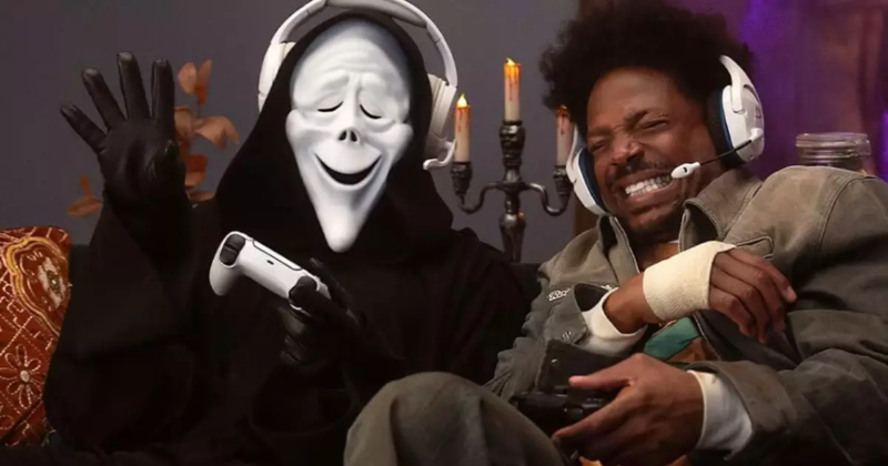 Cena de Todo Mundo em Pânico 6. Plano médio de duas figuras sentadas lado a lado em um sofá, jogando videogame. À esquerda, uma pessoa fantasiada como o vilão Ghostface, usando a clássica túnica preta, luvas e uma máscara branca sorridente modificada. Ela usa fones de ouvido brancos sobre a máscara, segura um controle de PlayStation 5 branco em uma das mãos e acena com a outra aberta. À direita, o ator Marlon Wayans aparece sorrindo e cerrando os dentes de forma cômica, usando fones de ouvido gamer brancos e uma jaqueta verde-oliva com uma faixa branca no punho, enquanto segura outro controle. Ao fundo, há um castiçal escuro com velas acesas.