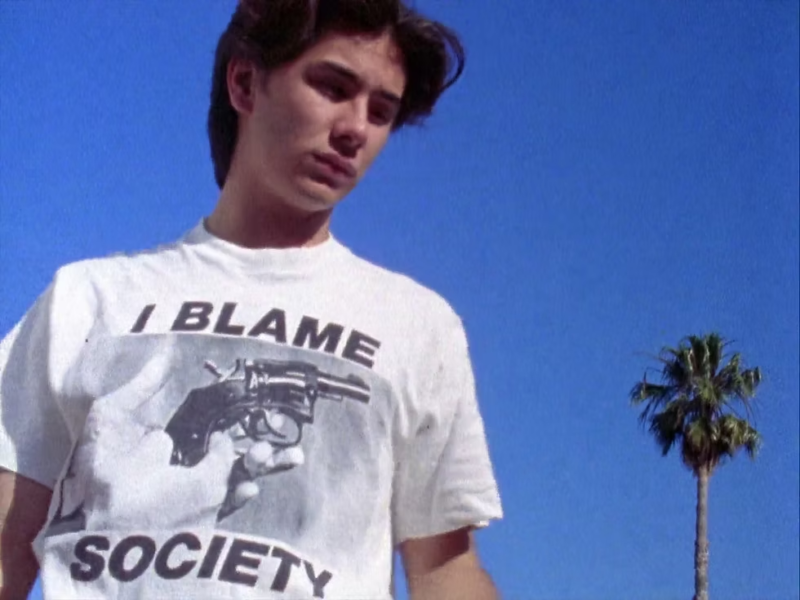 Cena do filme Totally Fucked Up. Na imagem, há um homem amarelo utilizando uma blusa branca com a estampa “Eu culpo a sociedade”. No meio da camiseta, há uma pessoa segurando uma arma.