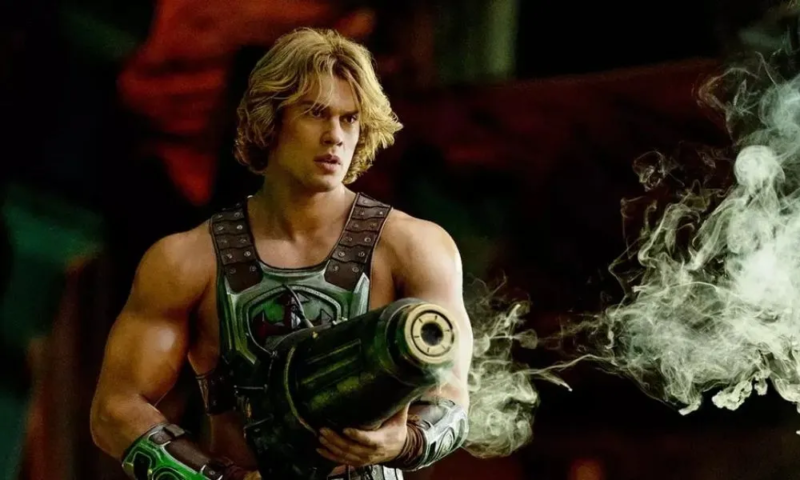 Cena do filme Mestres do Universo mostrando o He-Man, guerreiro loiro de porte atlético usando armadura metálica e segurando uma grande arma futurista. Ele observa algo fora de quadro com expressão séria e alerta, enquanto fumaça branca sai do cano da arma e se espalha pelo lado direito da imagem. O fundo escuro, iluminado por tons avermelhados e esverdeados, reforça a atmosfera de ação e conflito em um ambiente de fantasia e ficção científica.