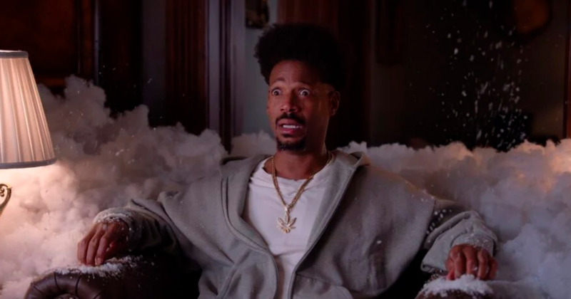 Cena de Todo Mundo em Pânico 6. Plano médio do ator Marlon Wayans sentado em uma poltrona de couro escuro, com uma expressão de extremo choque, olhos arregalados e boca aberta. Ele veste um casaco de moletom cinza aberto sobre uma camiseta branca e usa um colar de corrente dourada com um pingente em formato de folha de maconha. O personagem e toda a poltrona estão cercados e cobertos por uma grande quantidade de espuma branca densa ou neve artificial, que parece ter explodido no ambiente. À esquerda, vê-se a cúpula de um abajur aceso.