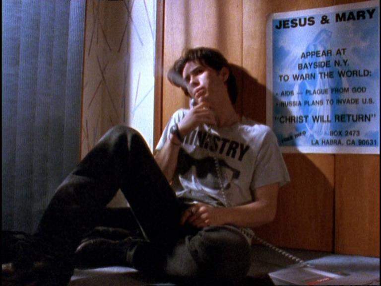 Cena do filme Totally Fucked Up. Na imagem, há um homem amarelo sentado no chão com um telefone fixo em mãos. Ele veste uma calça preta jeans e uma camiseta cinza. Atrás dele, à direita, há um cartaz com o título “Cristo retornará”.
