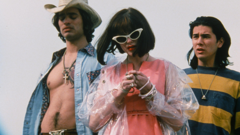 Três jovens estão lado a lado ao ar livre. No centro, uma mulher usa óculos de sol brancos, vestido rosa e uma capa transparente. À esquerda, um rapaz usa chapéu de cowboy e camisa aberta. À direita, outro rapaz veste uma camiseta listrada. Os três usam acessórios e roupas que remetem à moda alternativa dos anos 1990. 