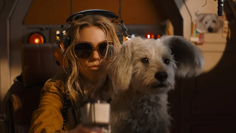 Cena do filme. À direita, um pequeno cachorro branco de pelos bagunçados, com uma das orelhas levantada. À esquerda, está Kara, jovem de cabelos loiros, usando óculos de sol grandes e redondos e fones de ouvido laranja. Ela tem uma expressão séria e assume o controle de uma cabine. O ambiente ao fundo é o interior de uma nave espacial.