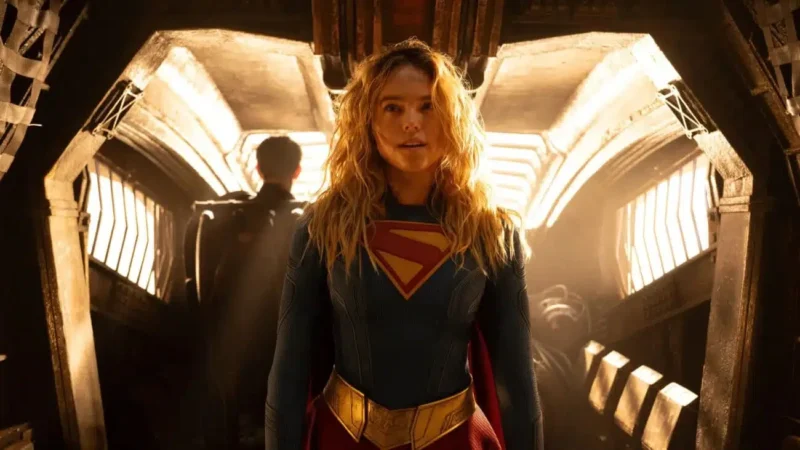  Kara, uma jovem mulher vestida com o uniforme de Supergirl, que é azul de manga longa com o símbolo vermelho e amarelo com 