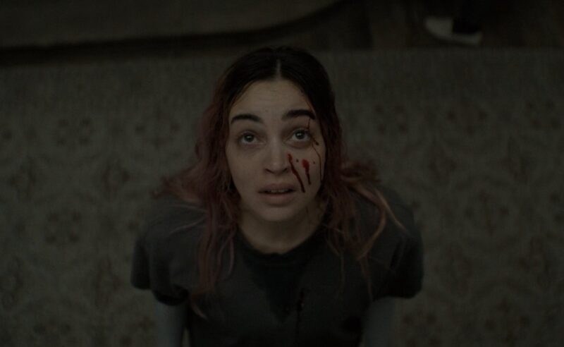Cena do filme A Morte do Demônio: Em Chamas Na imagem, vista de cima, Alice encara o teto. Na bochecha esquerda, há gotas de sangue escorrendo. Ela está com expressão de medo e veste uma camiseta cinza escuro com marcas de suor. Seu cabelo está desarrumado e possui mechas rosa. Alice é uma mulher na faixa dos 30 anos, de origem tunisiana e europeia.