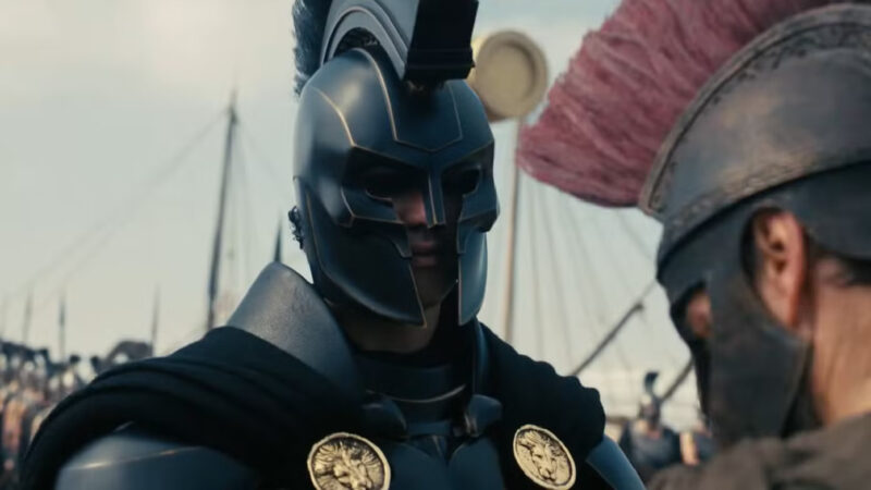 Dois guerreiros com armaduras de estilo grego antigo estão frente a frente. O da esquerda veste uma armadura preta fosca com detalhes dourados e capacete fechado, enquanto o da direita, visto de perfil, usa um capacete com uma grande pluma rosa. Ao fundo, há mastros de navios sob um céu claro.