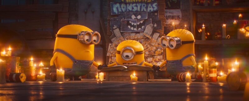 Cena do filme Minions e Monstros. Três minions estão em pé, cercados por velas acesas, em um ambiente que remete a um ritual. O minion do centro usa óculos de lente única e tem um livro aberto sobre uma caixa de madeira, com expressão séria. Ao fundo, um cartaz escrito "Minions y Monstras" mostra um rosto assustador e ilustrações de outros minions em pânico.