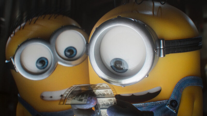 Cena do filme Minions e Monstros. Dois minions aparecem em close-up, em ambiente escuro. O da esquerda usa óculos de proteção com duas lentes redondas e tufos de cabelo escuro no topo da cabeça. O da direita usa um óculos com uma única lente grande presa por uma tira de couro. Os dois seguram, entre as mãos, um pedaço de filme fotográfico com quadros em preto e branco.