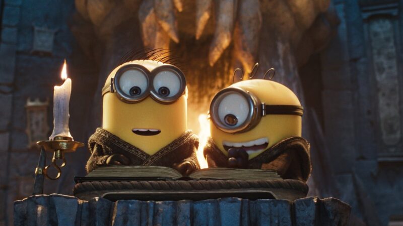Cena do filme Minions e Monstros. Dois minions vestem capas com capuz e aparecem atrás de um livro aberto sobre um púlpito de pedra, ao lado de uma vela acesa em castiçal dourado. O minion da esquerda usa óculos de duas lentes e tem mechas de cabelo espetadas. O da direita usa óculos de lente única e sorri, mostrando os dentes. Ao fundo, uma parede rochosa é iluminada por um brilho alaranjado.