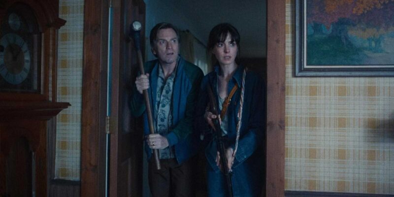 A imagem mostra um homem e uma mulher dentro de uma casa, posicionados na entrada de um cômodo. Os dois seguram objetos que parecem ser usados como armas improvisadas e observam o ambiente com expressão de alerta e apreensão. O homem veste uma jaqueta azul, enquanto a mulher usa uma jaqueta escura sobre uma camisa azul. Ao fundo, aparecem paredes revestidas com papel de parede xadrez, um quadro e parte de um móvel de madeira, criando uma atmosfera doméstica e sombria.