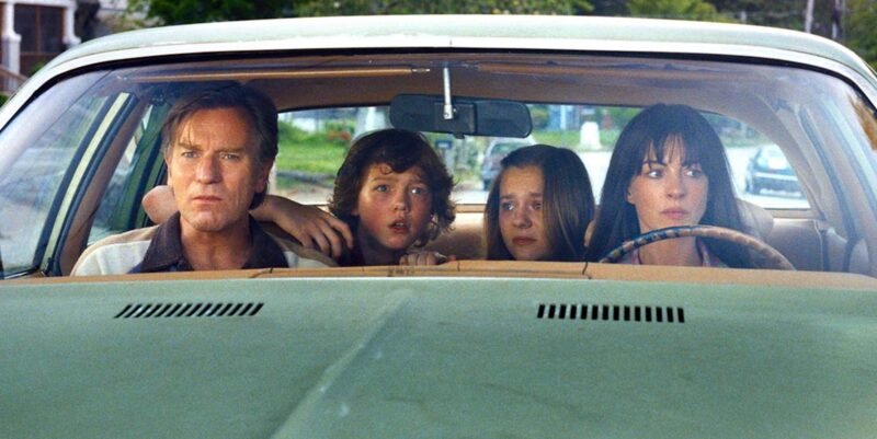 A imagem mostra quatro pessoas dentro de um carro antigo de cor verde-clara. No banco dianteiro, um homem dirige enquanto uma mulher está ao volante; entre eles, no banco traseiro, estão dois jovens. Os quatro observam atentamente o que acontece ao redor, com expressões de preocupação e tensão. A cena é registrada de frente, através do para-brisa do veículo, com uma paisagem residencial ao fundo.