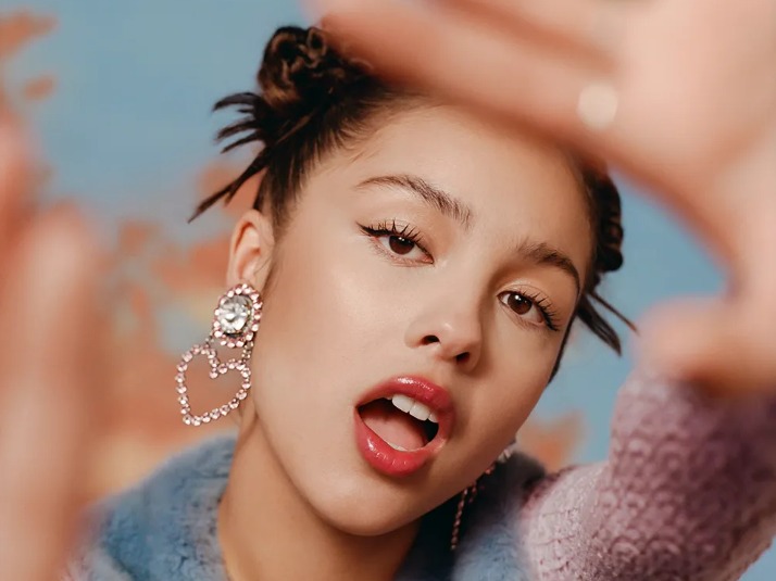  Foto da cantora Olivia Rodrigo, uma mulher amarela de cabelos ondulados. Na imagem, ela utiliza um penteado que prende seu cabelo. Ela usa um brinco de coração na orelha direita. Ela está com a boca aberta e com as mãos para frente.
