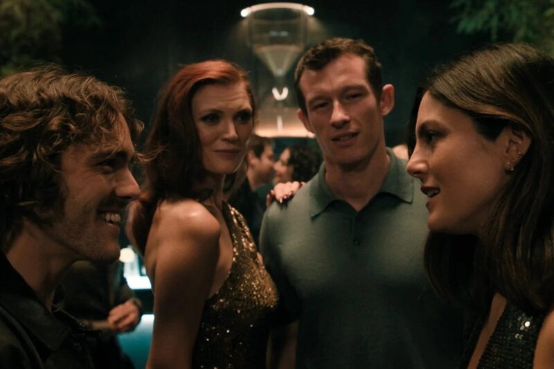 Cena noturna em um ambiente de festa, com quatro adultos conversando e sorrindo. À esquerda, um homem de cabelos castanhos cacheados veste uma jaqueta escura; ao centro, uma mulher ruiva usa um vestido preto brilhante ao lado de um homem de cabelo curto, que veste uma camisa verde. À direita, uma mulher de cabelos longos e castanhos, também com roupa escura e brilhante, participa da conversa.