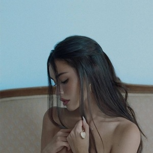 Capa do álbum locket de Madison Beer. A imagem mostra a cantora Madison Beer, uma mulher de pele branca e jovem. Ela aparece de perfil, com os olhos baixos e uma expressão melancólica. Seu cabelo escuro está preso em um penteado meio-preso, com mechas soltas emoldurando o rosto. Ela segura delicadamente um medalhão dourado em formato de coração contra o peito. O fundo apresenta uma parede azul clara e o topo de um sofá vintage bege.