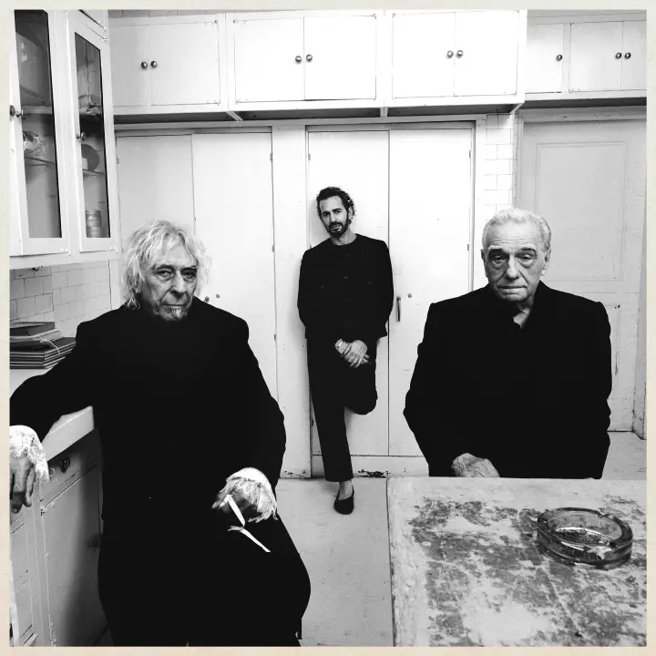 Texto alternativo: É uma foto em preto e branco tirada em uma cozinha com três homens: na esquerda está John Cale, um homem branco com cabelos e uma pequena barba branca, vestindo um casaco preto, sentado em um banco com a mão em uma bancada de cozinha. No meio, Marc Jacobs, homem branco com cabelo cacheado e barba, vestindo roupas pretas e uma sapatilha da mesma cor, com as mãos juntas encostado em um armário. Na esquerda, Martin Scorsese, homem branco com cabelos brancos e roupas pretas, sentado atrás de um balcão. Os três estão com uma expressão normal.