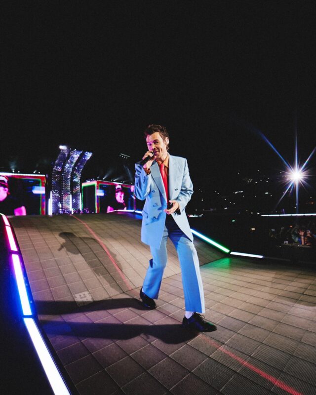 Harry Styles se apresenta no palco diante de uma grande plateia, segurando um microfone enquanto caminha por uma passarela iluminada. Vestindo um terno azul-claro com camisa e gravata em tons quentes, ele é o ponto central da composição. A iluminação do palco, os painéis de LED ao fundo e a escuridão do estádio destacam a dimensão do evento e a interação entre artista e público.