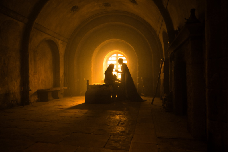 Cena do filme A Morte do Robin Hood. Plano geral de um vasto e antigo quarto com arquitetura de pedra, arcos e teto abobadado. A imagem é dominada por sombras profundas nas laterais, com o centro do cômodo iluminado dramaticamente por uma forte luz alaranjada que vem de uma janela em arco ao fundo. No centro, duas figuras estão em silhueta perfeita: uma pessoa deitada na cama e uma mulher em pé ao lado, parecendo oferecer algo ao deitado. A luz projeta uma sombra longa no chão de lajes de pedra. Há lareiras e bancos rústicos nas sombras.