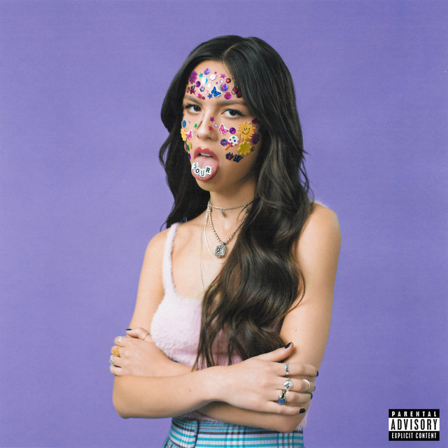 Capa do álbum Sour, da cantora Olivia Rodrigo. Na imagem, há a artista utilizando um top rosa felpudo com uma saia com listras branca, azul e roxa. O fundo da imagem é roxo e ela está no centro da capa. Ela possui cabelos ondulados e está com os braços cruzados. Ela mostra a língua, que contém 4 papéis que juntos formam a palavra Sour. Em seu rosto, há diversas figurinhas. No canto inferior direito, há o selo “Parental Advisory Explicit Content”. 