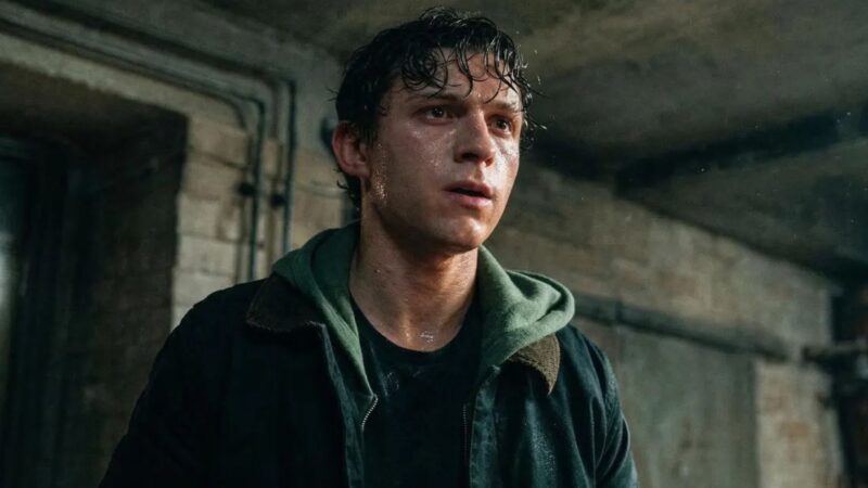 Cena do filme Homem-Aranha: Um Novo Dia. Na imagem, um jovem branco de cabelos castanhos curtos e molhados, com o rosto suado, veste um moletom verde com capuz sob uma jaqueta escura. Ele está em um ambiente com paredes de concreto ao fundo.