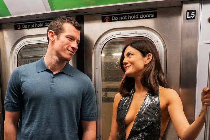 Duas pessoas estão em pé dentro de um vagão de metrô, diante das portas. Um homem de camisa polo azul olha sorrindo para uma mulher de cabelos longos e castanhos, que retribui o olhar enquanto segura uma barra de apoio. Ela veste um top preto brilhante de paetês, e ambos parecem estar se divertindo durante a viagem.