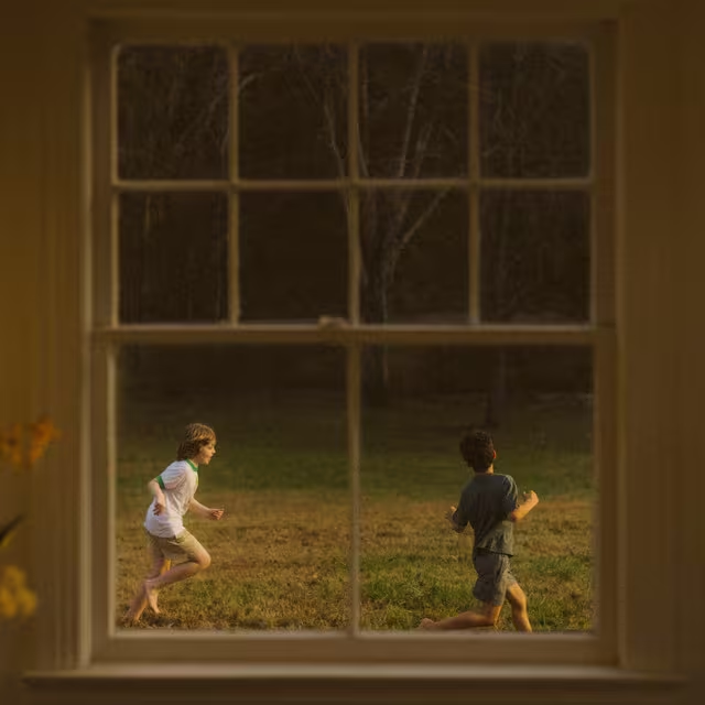 A imagem mostra a vista exterior de uma janela observada do lado de dentro da casa. As grades da janela e o entorno estão iluminados por uma luz amarelada. Do lado de fora da casa, dois meninos estão correndo em um gramado cercado por árvores secas. O menino à esquerda é loiro e usa uma camiseta branca e um shorts cinza, enquanto o menino à direita usa camiseta e shorts cinzas. Ambos estão brincando descalços.