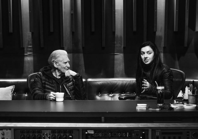 Texto Alternativo: Uma foto em preto e branco, na esquerda está David Cronenberg, homem branco com cabelos loiros e roupas pretas, está sentado olhando para Charli XCX com a mão no queixo e uma xícara na outra mão. Na direita, está Charli XCX, uma mulher branca com cabelos pretos e lisos, olhando para fora do ângulo, gesticulando a mão em um estúdio.
