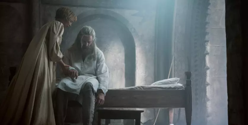 Cena do filme A Morte do Robin Hood. Plano médio de um cômodo rústico com paredes de pedra e arcos. Uma mulher de cabelo curto e vestido longo bege está de pé, segurando e observando atentamente a mão de um homem mais velho que está sentado na beira de uma cama de madeira. O homem tem longos cabelos grisalhos e barba, veste uma túnica branca e calças escuras, e olha para baixo. Uma luz azulada suave entra por uma abertura no alto da parede, criando uma atmosfera melancólica e de cuidado. Há um banco de madeira pequeno perto da cama.
