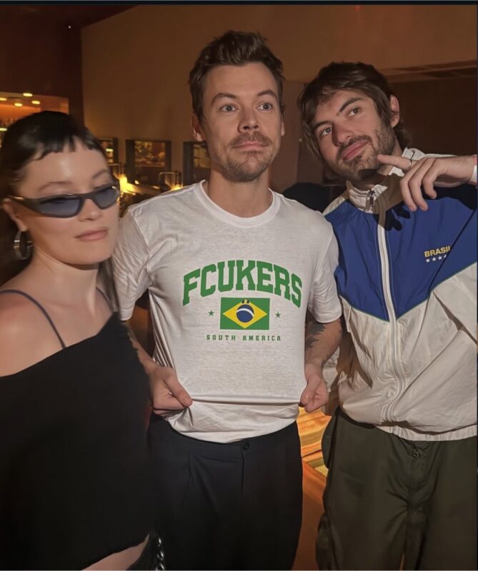 Três pessoas posam juntas em um ambiente interno após o evento. Ao centro, Harry veste uma camiseta branca com estampa em verde e amarelo e uma bandeira do Brasil, com o escrito ‘fcukers’, enquanto as pessoas ao seu lado aparecem em trajes casuais, uma delas usando óculos escuros e outra um agasalho esportivo. A fotografia transmite um clima descontraído de bastidores, registrando um momento de interação e proximidade após a apresentação.