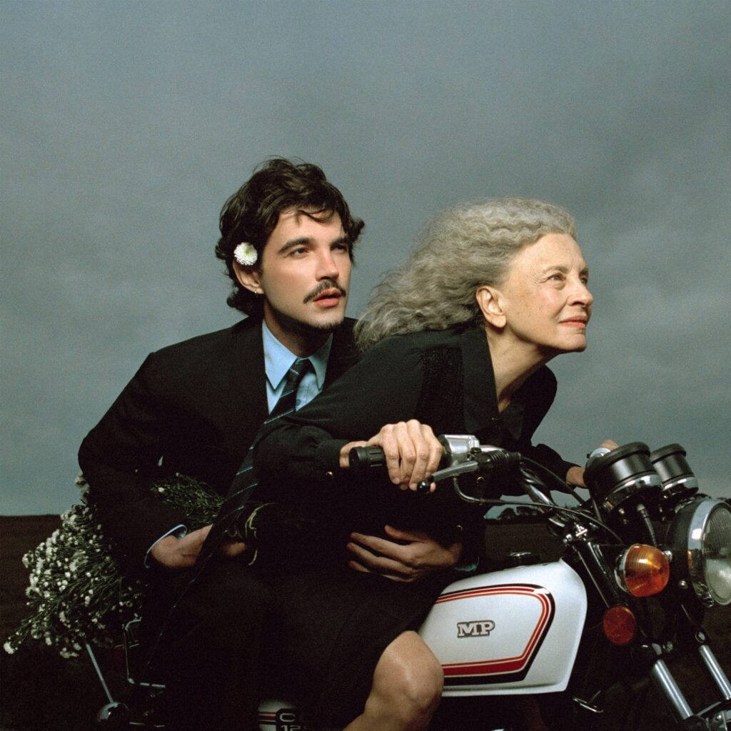 Texto alternativo: Capa do álbum Memórias Póstumas, do artista Jão, com a participação da atriz Marisa Sabino dos Santos. Uma mulher idosa de cabelos longos e grisalhos conduz uma motocicleta branca enquanto inclina o corpo para a frente e sorri discretamente, olhando para o horizonte. Atrás dela, um homem jovem de cabelos escuros e bigode a abraça pela cintura para se equilibrar. Ele veste terno preto com camisa azul e gravata, segura um buquê de pequenas flores brancas e tem uma flor presa ao cabelo acima da orelha esquerda. O céu ao fundo está completamente nublado, em tons de cinza, destacando a dupla e transmitindo sensação de liberdade e cumplicidade durante o passeio.