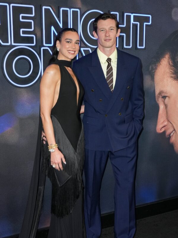 Duas pessoas posam lado a lado em um evento, diante de um painel iluminado com letras grandes e uma imagem de fundo. A mulher usa um vestido preto longo e elegante, com detalhes de franjas e acessórios dourados, enquanto o homem veste um terno azul-marinho, camisa clara e gravata listrada. Ambos sorriem para a câmera.