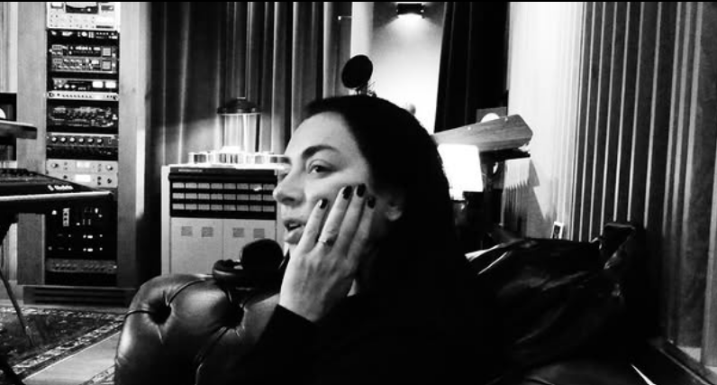 Texto Alternativo: Foto em preto em branco, Charli XCX, mulher branca com cabelos lisos e unhas escuras, está no foco da imagem olhando para o lado com a mão no rosto, sentada em um sofá no estúdio de música. 