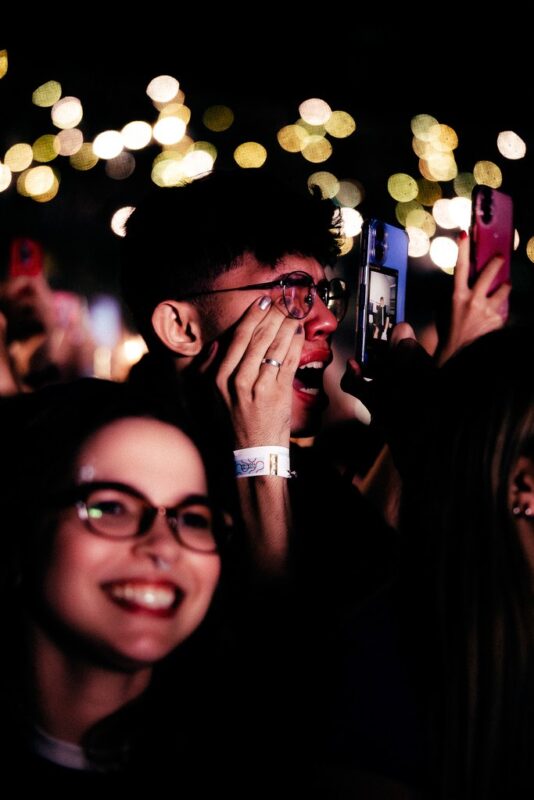 Um jovem na plateia reage de forma emocionada durante um show, levando as mãos ao rosto enquanto observa o palco e grava o momento com o celular. Ao redor, outros espectadores também utilizam seus aparelhos para registrar a apresentação. As luzes desfocadas ao fundo criam um efeito visual que evidencia a intensidade da experiência e o envolvimento do público com o espetáculo.