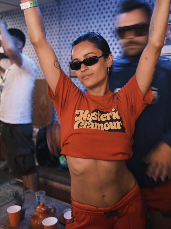 Uma mulher, Carla, usando óculos escuros e uma camiseta vermelha estampada, aparece com os braços erguidos durante um evento. A fotografia registra um momento de celebração e descontração, enquanto as pessoas ao fundo, incluindo Harry, logo atrás dela, aparecem desfocadas pelo movimento, reforçando a sensação de dinamismo e da atmosfera festiva do ambiente. O enquadramento destaca sua expressão serena em contraste com a energia ao redor.