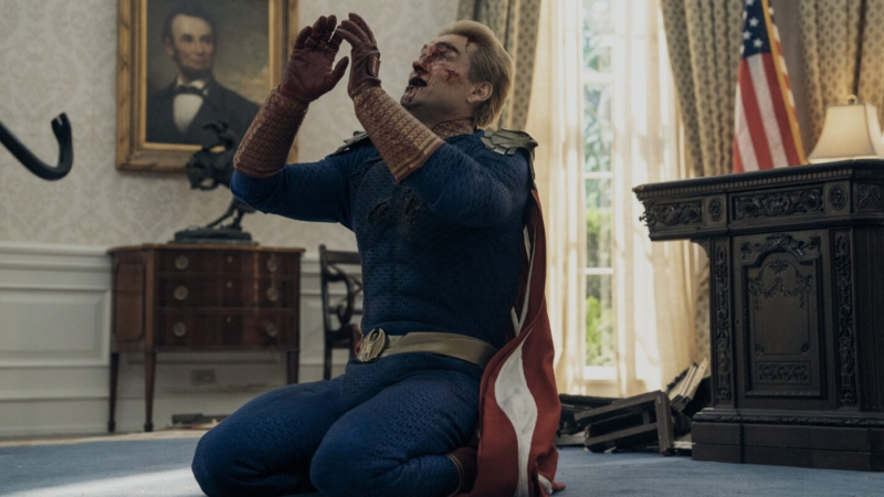 Cena de The Boys. Plano médio do Capitão Pátria ajoelhado no chão de carpete azul do Salão Oval. Ele veste seu traje de super-herói azul com detalhes dourados e capa vermelha. Seu rosto e cabelo estão visivelmente sujos de sangue, e ele está com a cabeça erguida e os olhos semicerrados, com as mãos erguidas em um gesto de desespero ou prece. Ao fundo, à esquerda, há um retrato emoldurado de Abraham Lincoln na parede e, à direita, parte de uma mesa de madeira escura e a bandeira dos Estados Unidos.