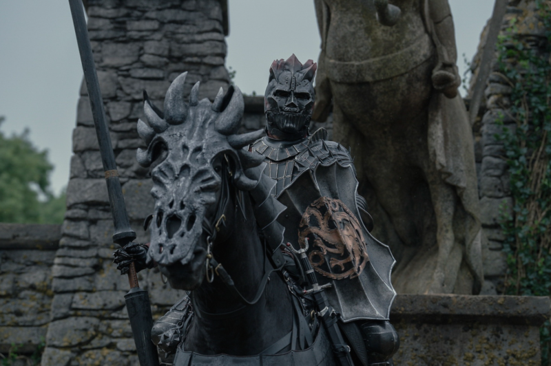 Cena da série O Cavaleiro dos Sete Reinos. Um cavaleiro imponente veste uma armadura negra ornamentada e segura uma lança enquanto monta um cavalo igualmente protegido por uma armadura com formato de cabeça de dragão. O elmo do cavaleiro possui detalhes pontiagudos e fecha completamente o rosto. No peito, um escudo com o símbolo de um dragão em tons avermelhados se destaca. Ao fundo, uma construção de pedra e uma estátua parcialmente visível reforçam o ambiente medieval.