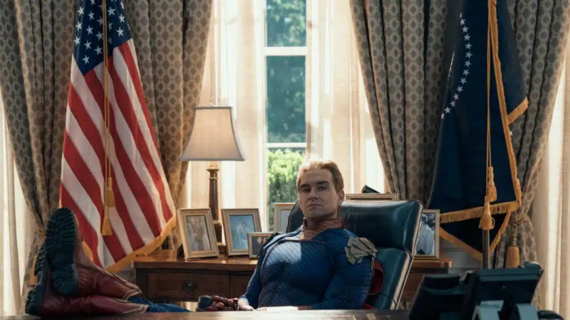 Cena de The Boys. O Capitão Pátria sentado de forma imponente e relaxada na cadeira presidencial do Salão Oval, atrás de uma grande mesa de madeira. Ele está com os pés calçados com suas botas vermelhas apoiados sobre a mesa, cruzando as pernas. Seu semblante é sério, frio e desafiador, olhando diretamente para a câmera. Ao fundo, janelas iluminadas estão emolduradas por cortinas pesadas com padrões geométricos, ladeadas pela bandeira americana à esquerda e pela bandeira presidencial azul à direita.
