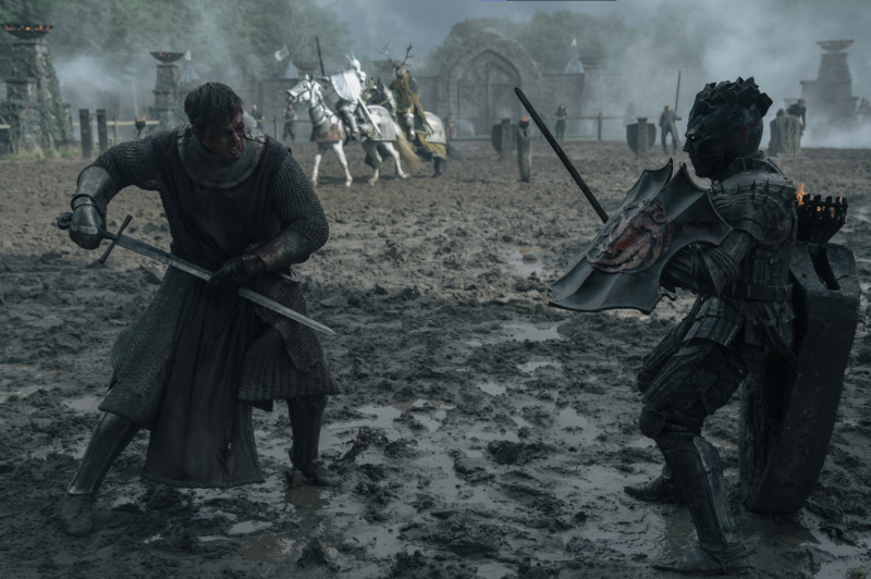 Cena da série O Cavaleiro dos Sete Reinos. Na imagem, dois guerreiros se enfrentam em um campo de batalha enlameado e nebuloso. À esquerda, um homem vestido com cota de malha e túnica cinza empunha uma espada com as duas mãos, em postura de ataque, com o rosto tenso e sujo. À direita, uma figura coberta por uma armadura escura e imponente segura um escudo com detalhes em vermelho e uma espada. Ao fundo, cavaleiros montados em cavalos brancos avançam em meio à névoa, próximos a um arco de pedra.