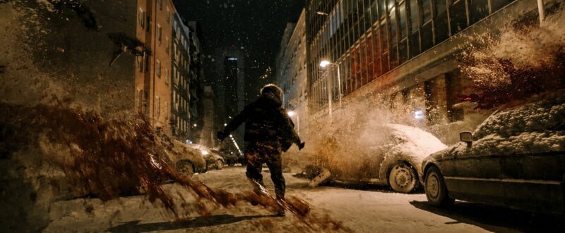 Cena do filme Resident EvilNa imagem, o personagem Bryan, de costas, corre por uma rua com carros abandonados e cobertos de neve enquanto pessoas se atiram do prédio, explodindo em sangue no chão e nos automóveis. O cenário é noturno, há edifícios e iluminação oriunda dos postes. Há neve por toda parte e Bryan veste calça e jaqueta escura.